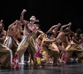 Diversos bailarinos com figurinos coloridos levantando os braços em conjunto.