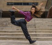 Bailarina com roupa roxa e calças pretas em um salto na frente das escadarias da Praça das Artes