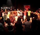 Grupo de pessoas estão lado a lado com as mãoes dadas e levantadas em um circulo