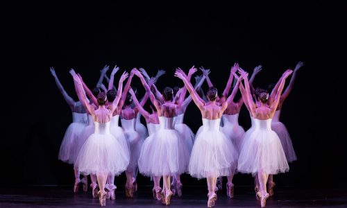 Bailarinas vestidas de branco se apresentando em um cenário escuro, com fundo preto.