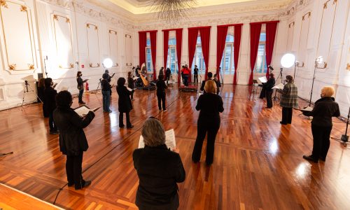 6_Coral Paulistano_Foto_Rafael_Salvador Integrantes do Coral Paulistano com roupas pretas e segurando partituras, dispostos na sala do conservatório. Luzes estão dispostas na sala e o ambiente esta bem iluminado e sem cadeiras.