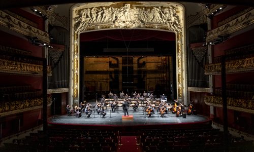 Orquestra-Sinfônica-Municipal-SP_RafaelSalvador159 Sala de Espetáculos do Theatro Municipal com o palco iluminado no centro. No palco estão os membros e o regente da Orquestra Sinfônica Municipal. Nas laterais e estão três andares de poltronas e na parte inferior algumas pessoas assistindo.
